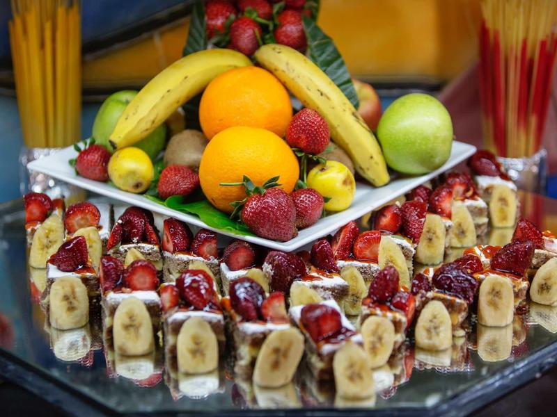 Vassoio colorato di frutta e dessert con banane, fragole, arance e uva disposti.