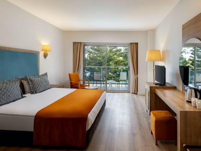 Modern hotelkamer met tweepersoonsbed, bureau, tv en balkon met uitzicht op bomen.