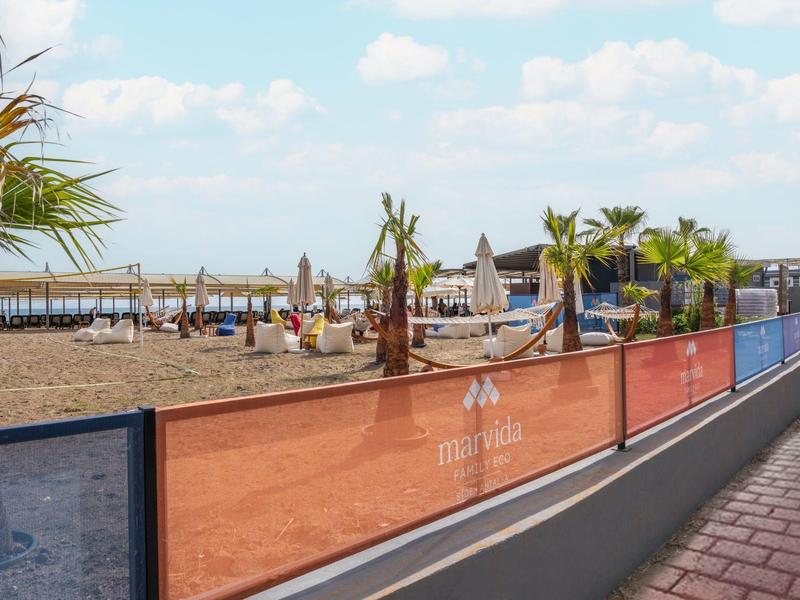 Playa con palmeras, sombrillas y turistas, bordeada por una cerca junto a un camino peatonal.