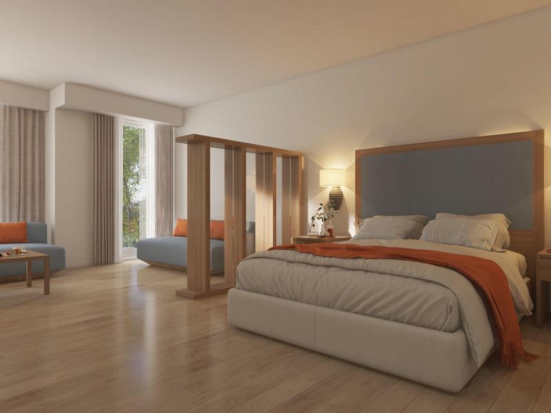 Habitación moderna de hotel con cama grande, zona de estar y luz natural a través de grandes ventanas.