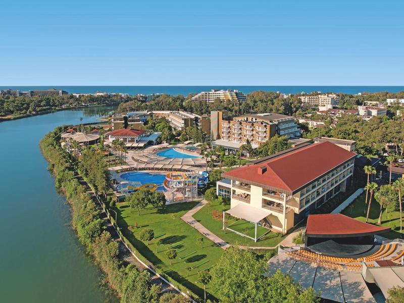 Complejo hotelero junto a un río con varios edificios, piscinas y áreas verdes bajo un cielo azul.