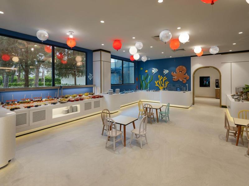 Área infantil luminosa y colorida con mesas, sillas y decoraciones en las paredes bajo luces de colores.