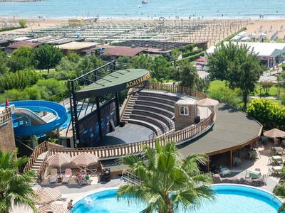 Freizeitpark mit blauen Wasserrutschen, runder Bühne, Pool und Sandstrand im Hintergrund.