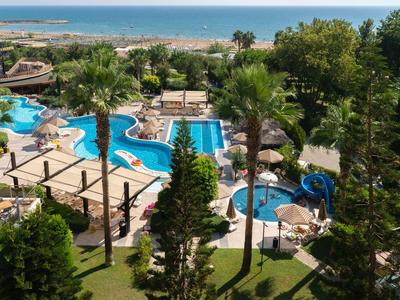 Blick auf einen Hotelpool mit Palmen, blauen Wasserrutschen und dahinter das Meer am Horizont.