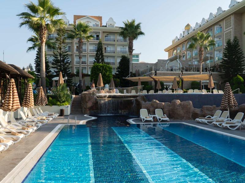 Duży basen zewnętrzny w hotelu z leżakami i palmami w słoneczny dzień.