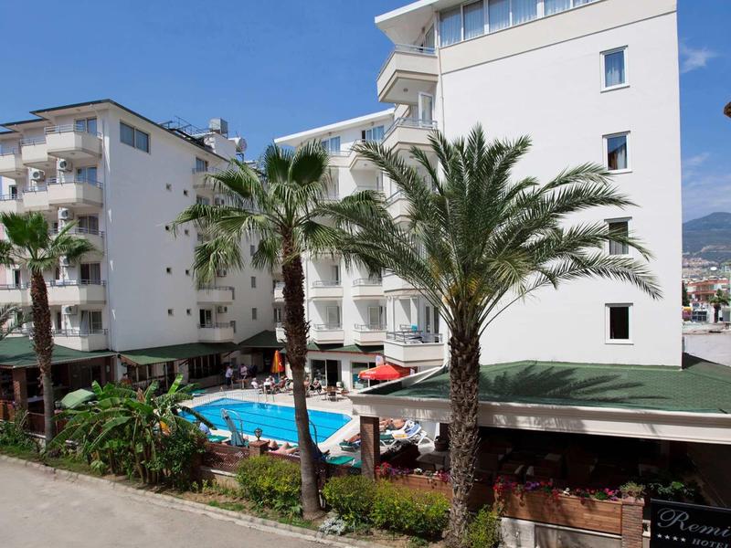 Hôtel avec piscine extérieure, plusieurs palmiers et jardin soigné sous un ciel bleu.