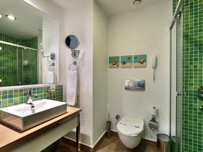 Modernes Badezimmer mit grün gefliester Dusche, weißem Waschbecken und WC, helle Wände und Holzboden.