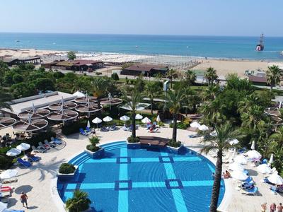 Large hotel pool with umbrellas and loungers, palm trees, and a view of the beach and sea.