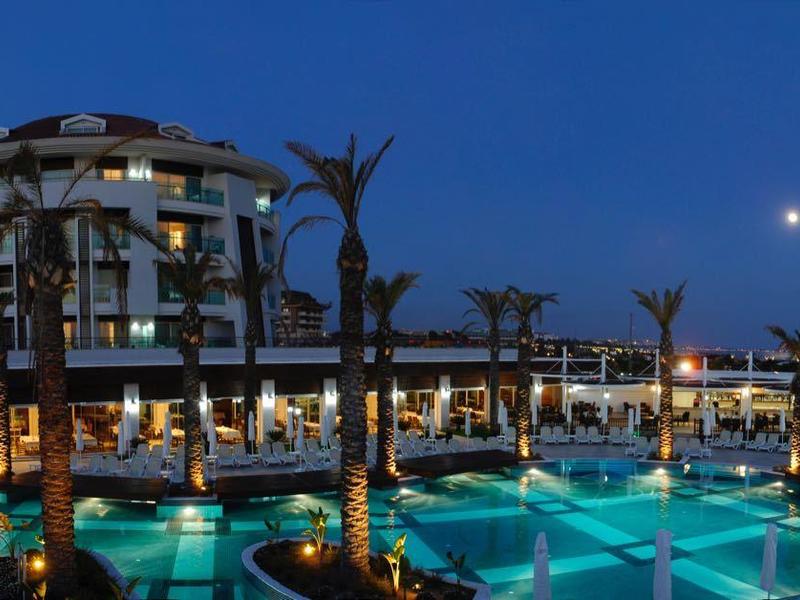 Großes Hotel mit beleuchteten Pools und Palmen, umgeben von Lichtern in der Abenddämmerung.