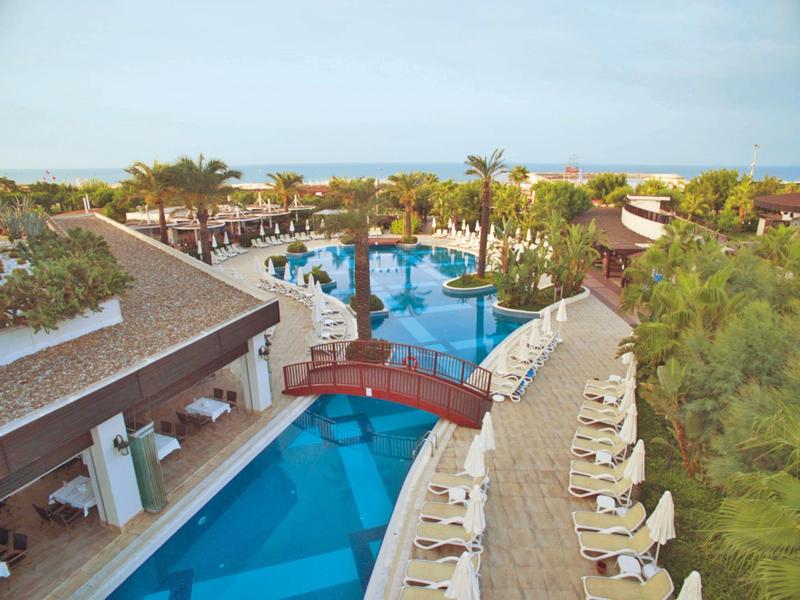 Großer blauer Pool mit Brücke, Palmen und Liegestühlen in einem tropischen Resort.