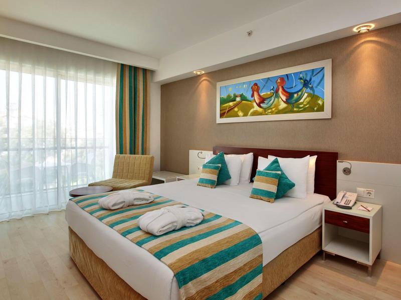 Modernes Hotelzimmer mit großem Bett, bunter Bettwäsche, Nachttisch und Wandbild mit Hühnermotiven.