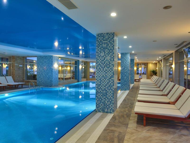 Innenpool mit blauen Kacheln, Liegestühlen mit weißen Polstern und großen Fenstern.