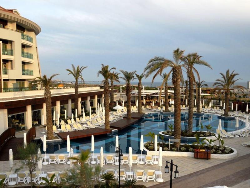 Hotelpool mit Palmen, Sonnenliegen und mehrstöckigem Gebäude bei bewölktem Himmel.