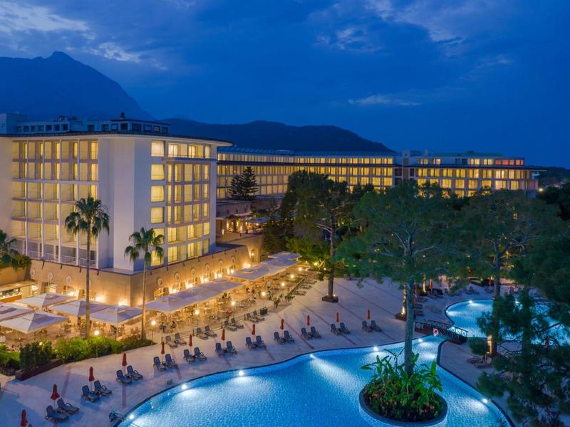 Elegancki hotel z oświetlonymi basenami i górami w tle o zmierzchu.