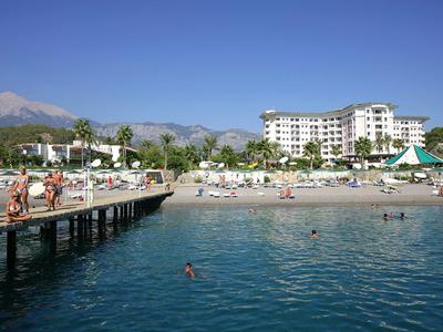 Spiaggia con molo di legno, ombrelloni, hotel e montagne sullo sfondo sotto un cielo limpido.