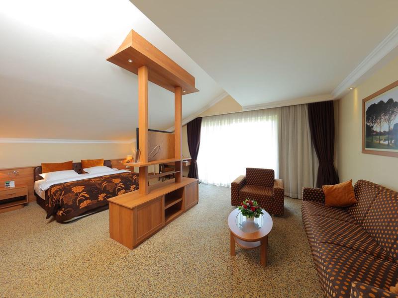 Prostorný hotelový pokoj s manželskou postelí, pohovkou, křeslem a velkým oknem se závěsy.