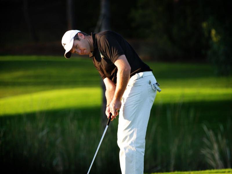 Golfista v bílých kalhotách a čepici odpaluje míček na dobře udržovaném golfovém hřišti.