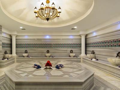 Grande hammam dell'hotel con bacino in marmo e pareti decorative, illuminato da un lampadario.