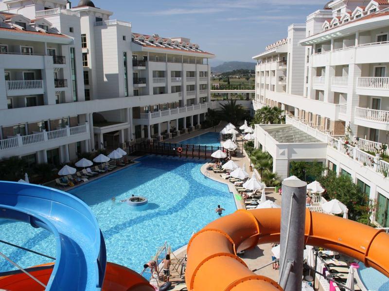 Velký venkovní bazén s modrými a oranžovými skluzavkami mezi hotelovými budovami.