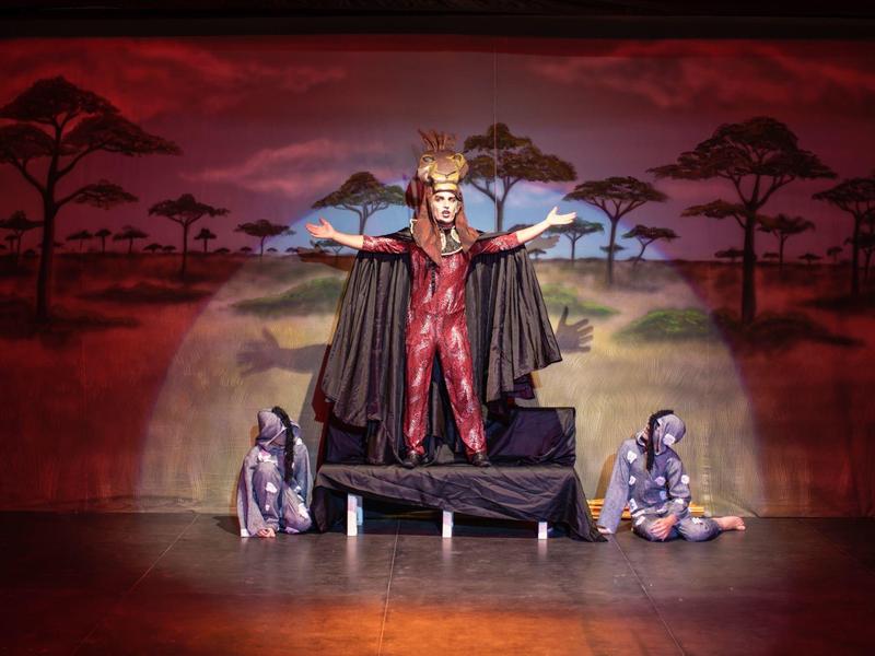 Divadelní scéna s královnou v červených šatech a plášti, dvě postavy ve stříbrných kostýmech sedí vlevo a vpravo.