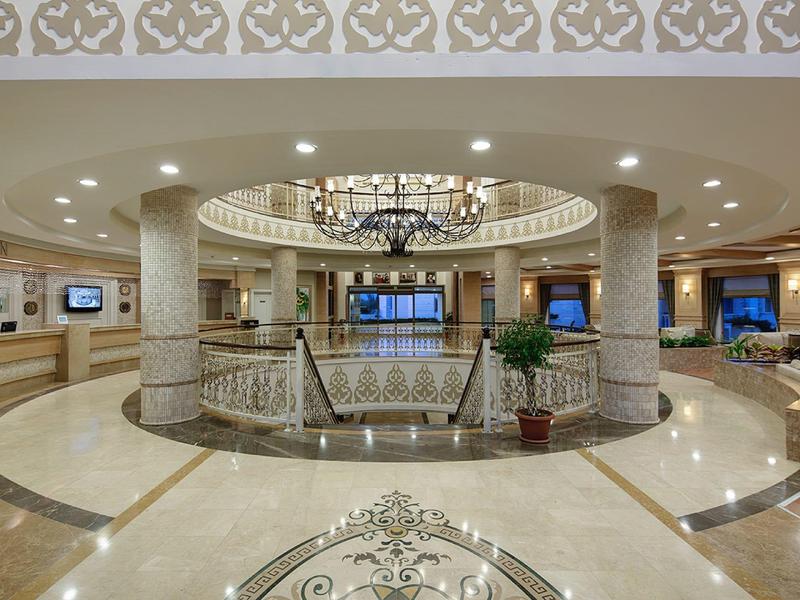 Velká hotelová hala se schodištěm, mramorovou podlahou a elegantním lustrem.