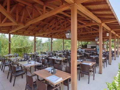Offene Holzterrasse mit Tischen und Stühlen, umgeben von Bäumen, unter blauem Himmel.