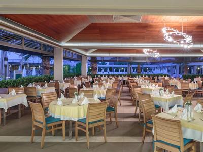 Großes, offenes Restaurant mit hellen Tischen, Holzstühlen und moderner Deckenbeleuchtung bei Abenddämmerung.