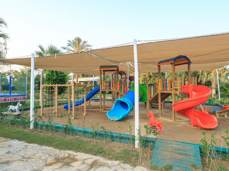 Spielplatz mit bunten Rutschen und Klettergeräten unter einem Sonnensegel in einem Hotelgarten.