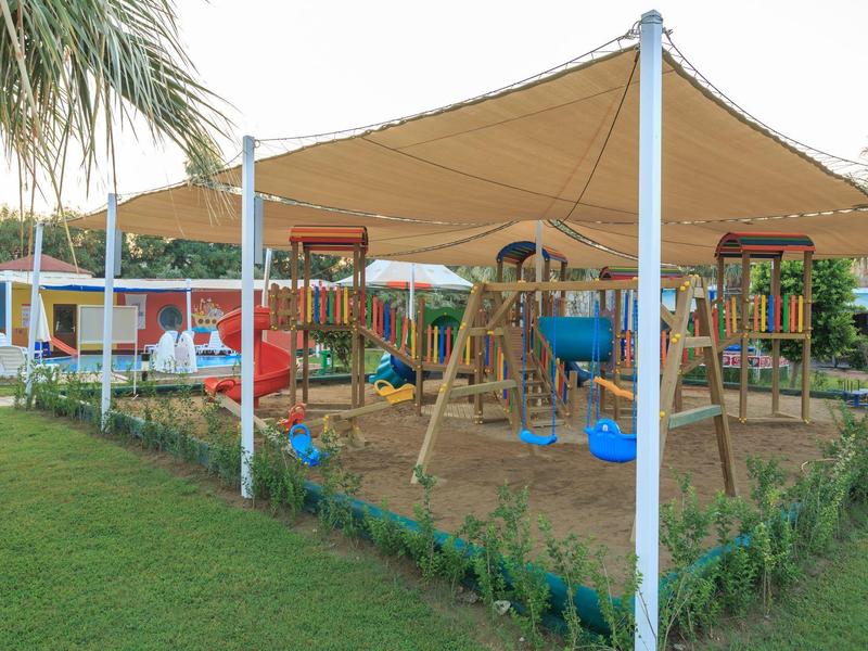 Überdachter Kinderspielplatz mit Rutsche, Schaukeln und Sandfläche im Freien.