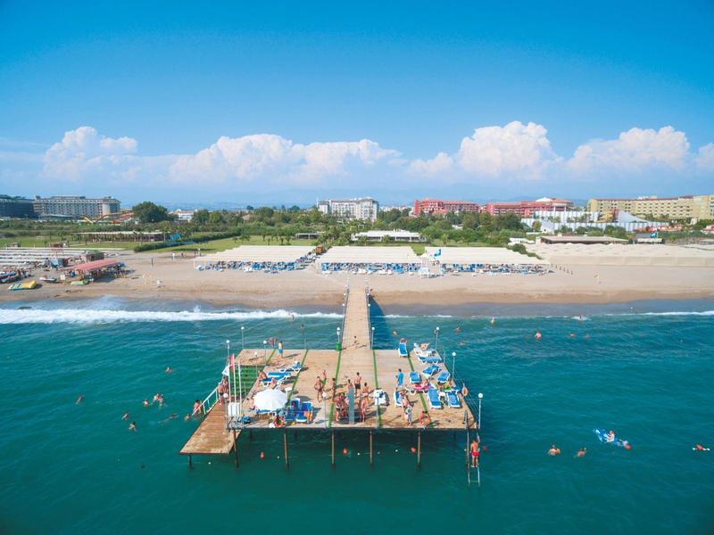 Langer Holzsteg führt von einem Sandstrand mit Liegestühlen ins blaue Meer unter blauem Himmel.