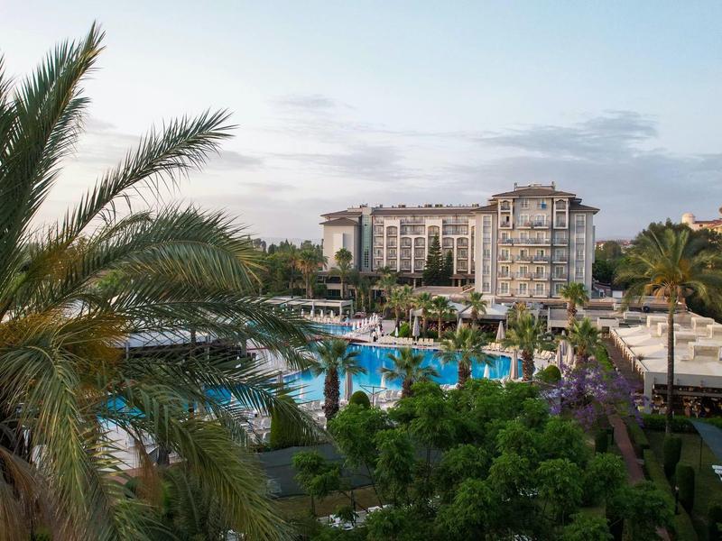 Große Hotelanlage mit Pool, Palmen und grünen Büschen, blauer Himmel im Hintergrund.