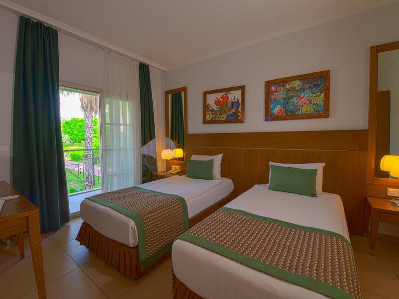 Zwei Einzelbetten in schlichtem Zimmer mit Holzmöbeln, grünen Vorhängen und bunten Bildern an der Wand.