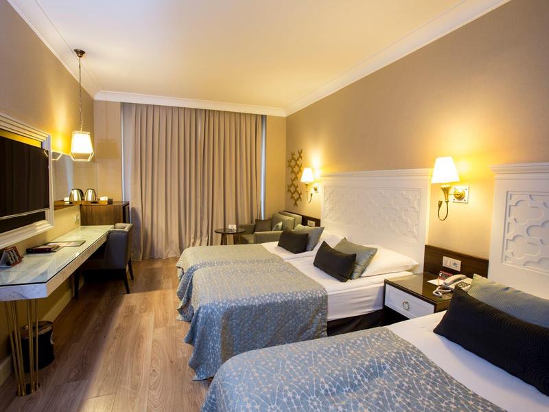 Camera d'hotel luminosa con due letti, scrivania, TV a schermo piatto e tenda alla finestra.