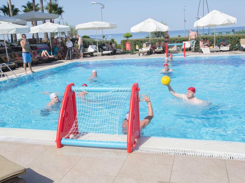 Partita di pallanuoto in piscina all'aperto di un hotel in una giornata di sole con giocatori e spettatori.