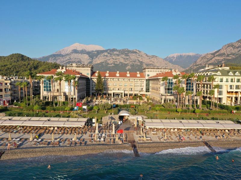Hotel e edifici lungo la costa con montagne sullo sfondo e una spiaggia di ciottoli in primo piano.
