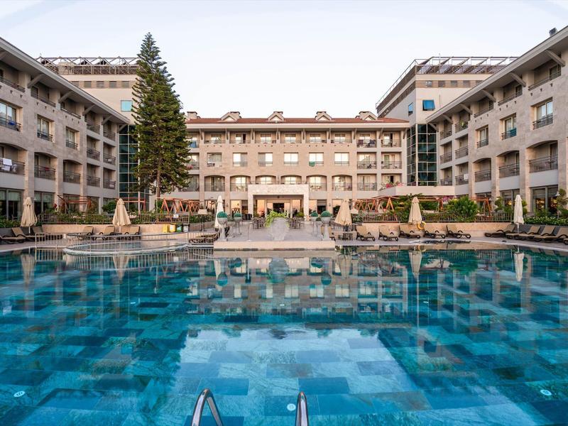 Hotel di lusso con grande piscina all'aperto, circondato da edifici a più piani e ombrelloni.
