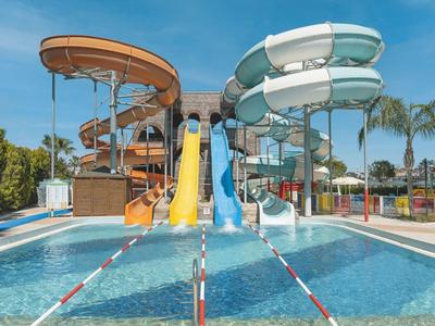 Mehrere bunte Wasserrutschen münden in einen Pool, umgeben von Palmen unter blauem Himmel.