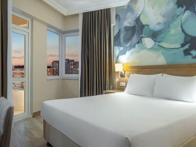 Moderní hotelový pokoj s manželskou postelí, psacím stolem a velkým oknem s výhledem na město.