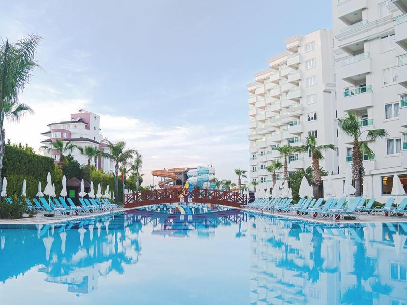 Velký čistý bazén s lehátky a palmami vedle moderního bílého hotelu.