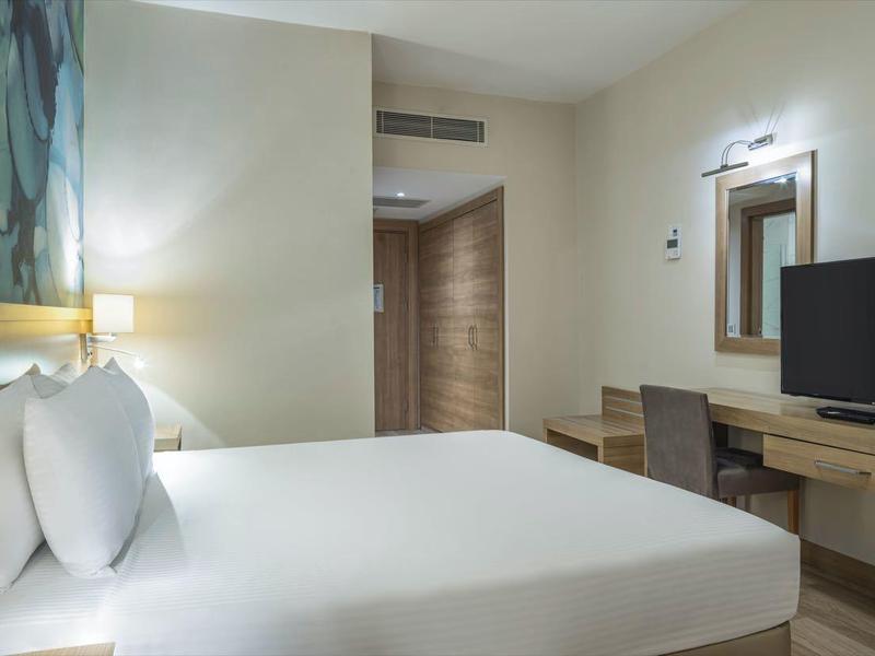 Moderní hotelový pokoj s manželskou postelí, televizí a světlým dřevěným stolem.