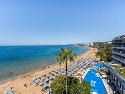 Una amplia playa de arena con tumbonas y palmeras junto a un hotel con piscina y vista al mar azul.