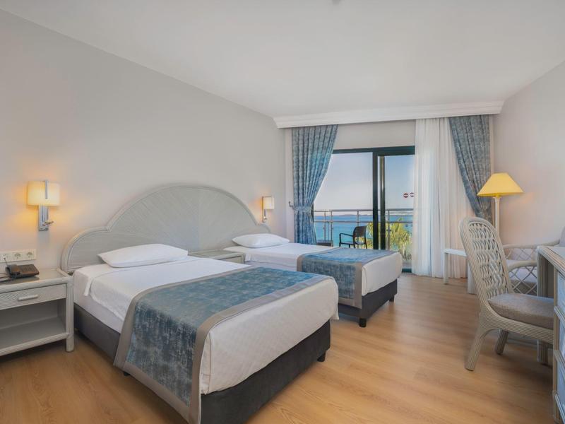 Habitación de hotel con dos camas individuales, suelo de madera, escritorio y balcón con vista al mar.