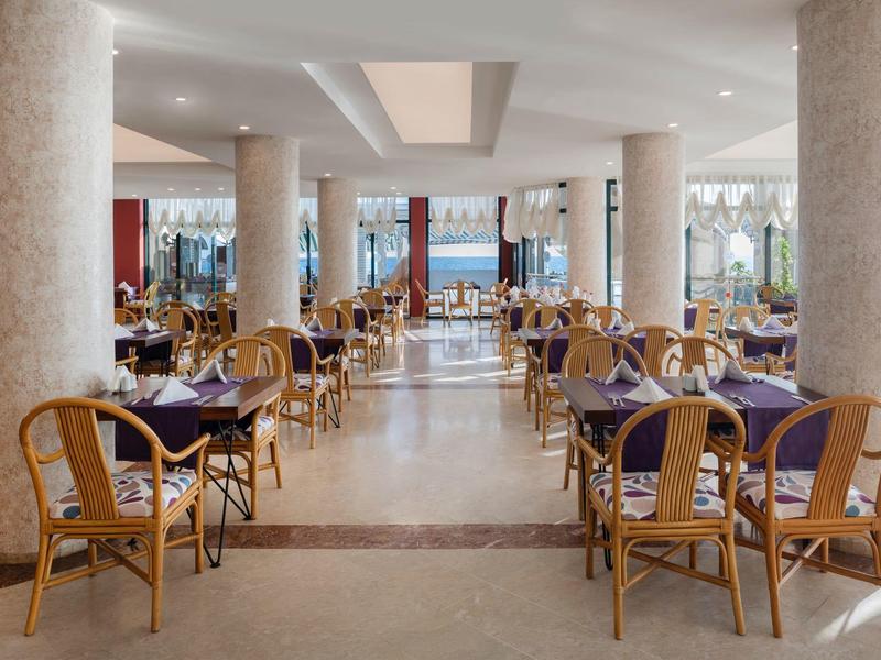 Amplio comedor de hotel con sillas de bambú y mesas con manteles oscuros, piso claro y columnas.