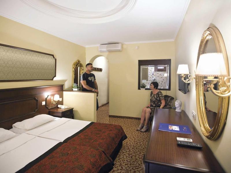 Chambre d'hôtel avec lit double, bureau, miroir et deux personnes, une près du lit et une assise sur une chaise.