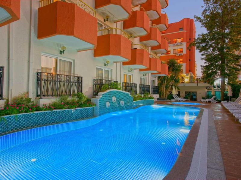 Hôtel avec piscine longue, entrée voûtée et balcons rouges sous un ciel clair.