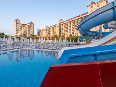 Großer Hotelpool mit Wasserrutschen und vielen weißen Liegestühlen vor einem mehrstöckigen Hotelgebäude.