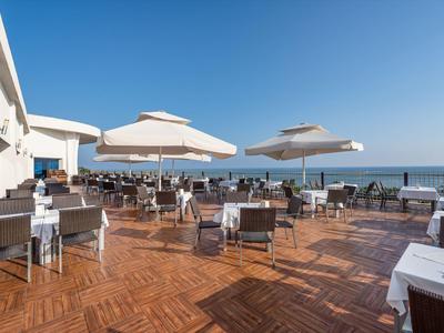 Velká hotelová terasa s stoly, židlemi a slunečníky s výhledem na moře.