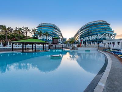 Moderní hotelový komplex s zakřivenými skleněnými budovami a velkým venkovním bazénem při západu slunce.