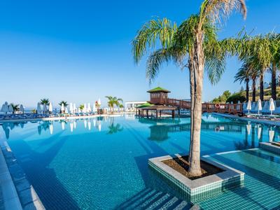 Velký hotelový bazén s palmami a výhledem na moře pod jasnou modrou oblohou.