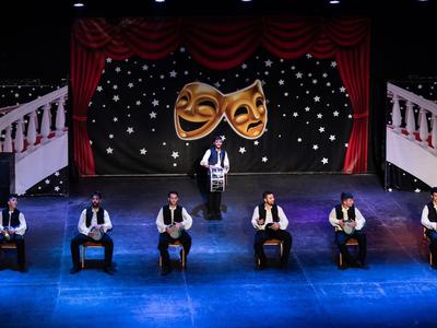 Sei uomini in abiti tradizionali eseguono una danza su un palco con maschere teatrali e tende.